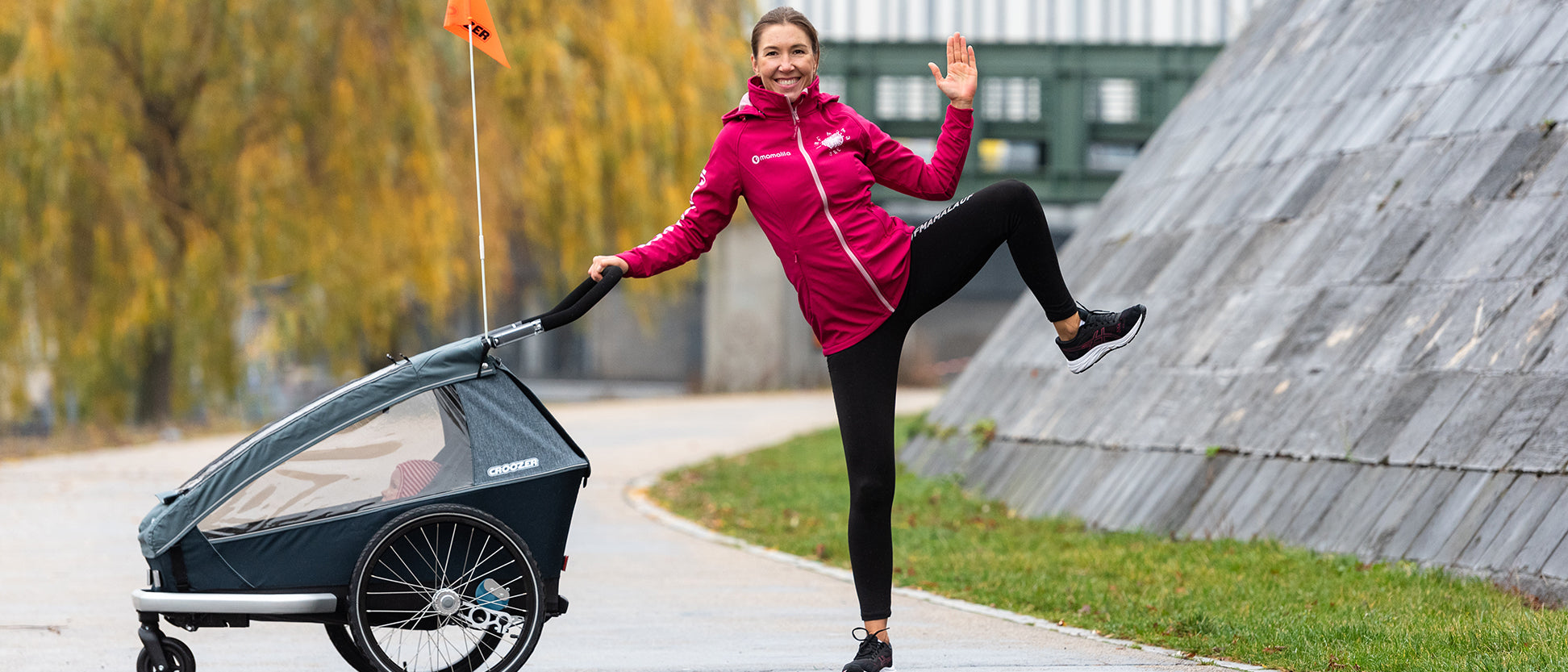 Smiling woman with on one leg and hand up while holding Croozer bike trailer with autumn backdrop in city park